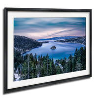 A framed photograph depicting a serene landscape with a lake surrounded by a forested area and mountains in the distance.