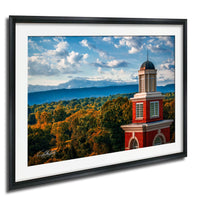A framed image depicting a red brick building with a dome, surrounded by a lush green forest and a blue sky with clouds.