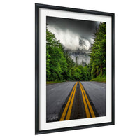 a winding road with yellow double lines, surrounded by lush green trees and a dramatic cloudy sky in the background.