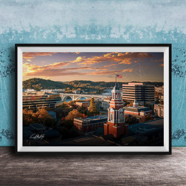 The image is a framed photograph depicting a cityscape with a bridge, buildings, and a clock tower, all set against a colorful sunset sky.