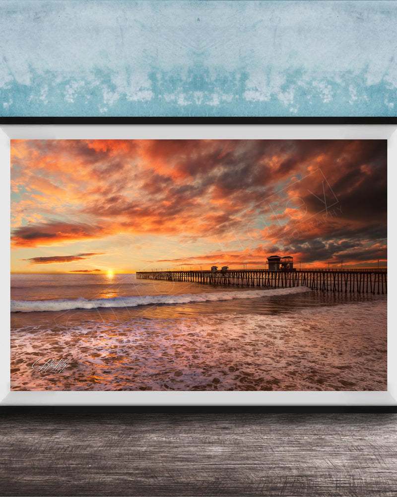 A framed photograph depicting a serene beach scene at sunset, with a wooden pier extending into the water and the sky filled with vibrant orange and pink hues.