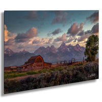 A rustic wooden barn sits in a grassy field, with majestic mountains rising in the background under a colorful sky at sunset.