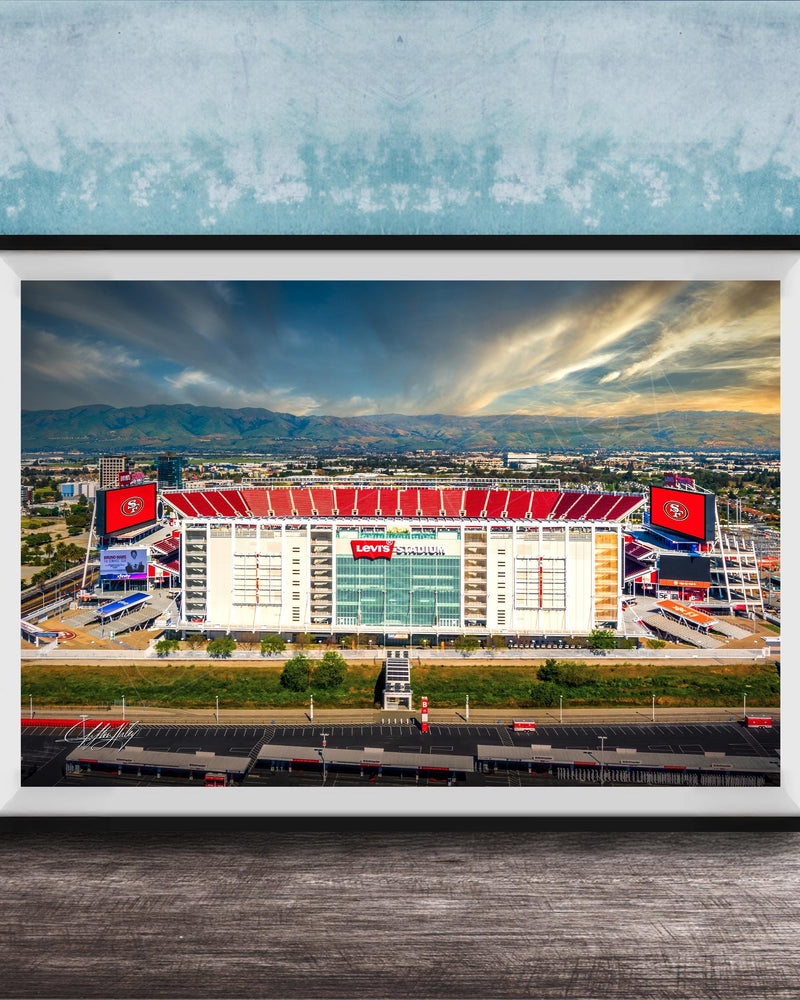 A framed photograph of a large sports stadium with a red roof, surrounded by mountains and a cloudy sky.