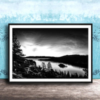 A black and white landscape photograph depicting a serene lake surrounded by mountains and pine trees, with a cloudy sky overhead.