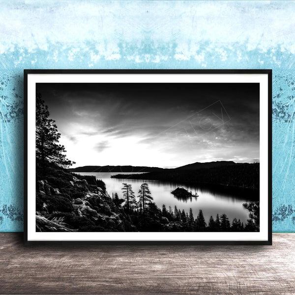 A black and white landscape photograph depicting a serene lake surrounded by mountains and pine trees, with a cloudy sky overhead.