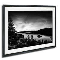 A black and white landscape photograph depicting a serene lake surrounded by mountains and trees, with a small island in the center.
