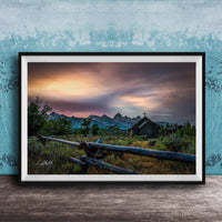 A framed photograph depicting a serene rural landscape with a wooden fence, a church, and mountains in the distance, set against a colorful sunset sky.