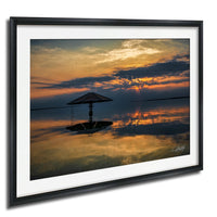 A framed photograph depicting a serene sunset over a calm body of water, with a silhouetted umbrella in the foreground.