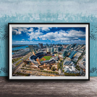 The image is an aerial view of a city with a baseball stadium in the center, surrounded by tall buildings and a body of water.