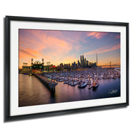 A framed photograph depicting a city skyline at sunset, with boats docked in a harbor and a large stadium in the background.