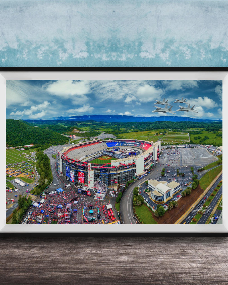 The image is an aerial view of a large stadium surrounded by a green landscape, with a few buildings and vehicles visible.