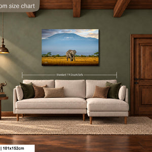 a living room with a large painting of an elephant in a field hanging on the wall. The painting is framed and measures approximately 40 inches by 101.5 centimeters. The room has a wooden floor and a beige sofa with throw pillows.