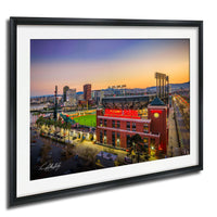 A framed aerial view of a city skyline at sunset, with a baseball stadium and surrounding buildings illuminated by colorful lights.