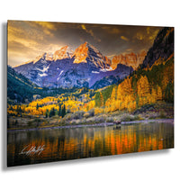 A breathtaking mountain landscape with a lake in the foreground, surrounded by vibrant fall foliage. The mountains are covered in snow and their peaks are bathed in warm, golden light, creating a stunning contrast against the cloudy sky. A moose can be seen in the distance, adding to the serene and picturesque scene.