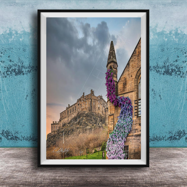 A framed photograph of a castle with a tower, set against a cloudy sky, with a colorful flower arrangement hanging from the tower.