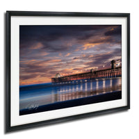 A framed photograph depicting a serene coastal scene at sunset, with a wooden pier extending into the calm waters and the sky filled with vibrant hues of orange and pink.