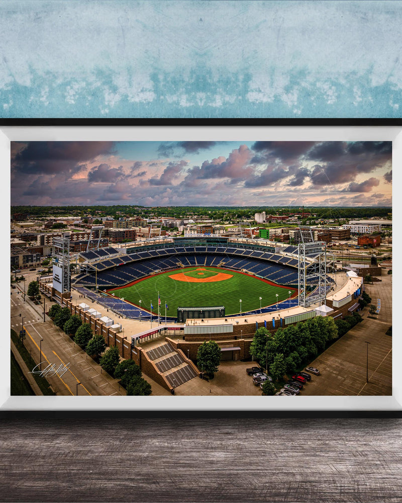 An aerial view of a baseball stadium with a lush green field, surrounded by buildings and a cloudy sky.