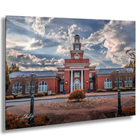 A brick building with a prominent clock tower, surrounded by a landscaped area with trees and bushes. The sky is filled with clouds, creating a dramatic backdrop for the scene.