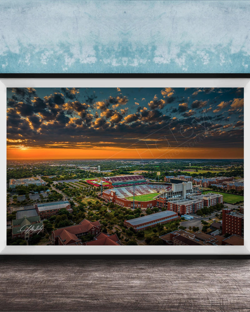 The image is a framed aerial view of a cityscape at sunset, with the sky filled with clouds and the sun setting over the horizon.