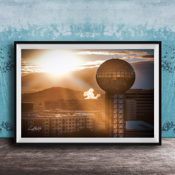 A framed photograph of a city skyline at sunset, with a large tower in the foreground and the sun setting behind the buildings.