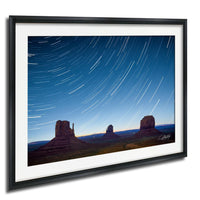 A framed photograph depicting the iconic rock formations of Monument Valley at night, with star trails visible in the sky.