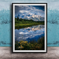 A framed photograph of a serene landscape with a mountain range reflected in a calm lake, surrounded by lush greenery and vibrant yellow flowers.