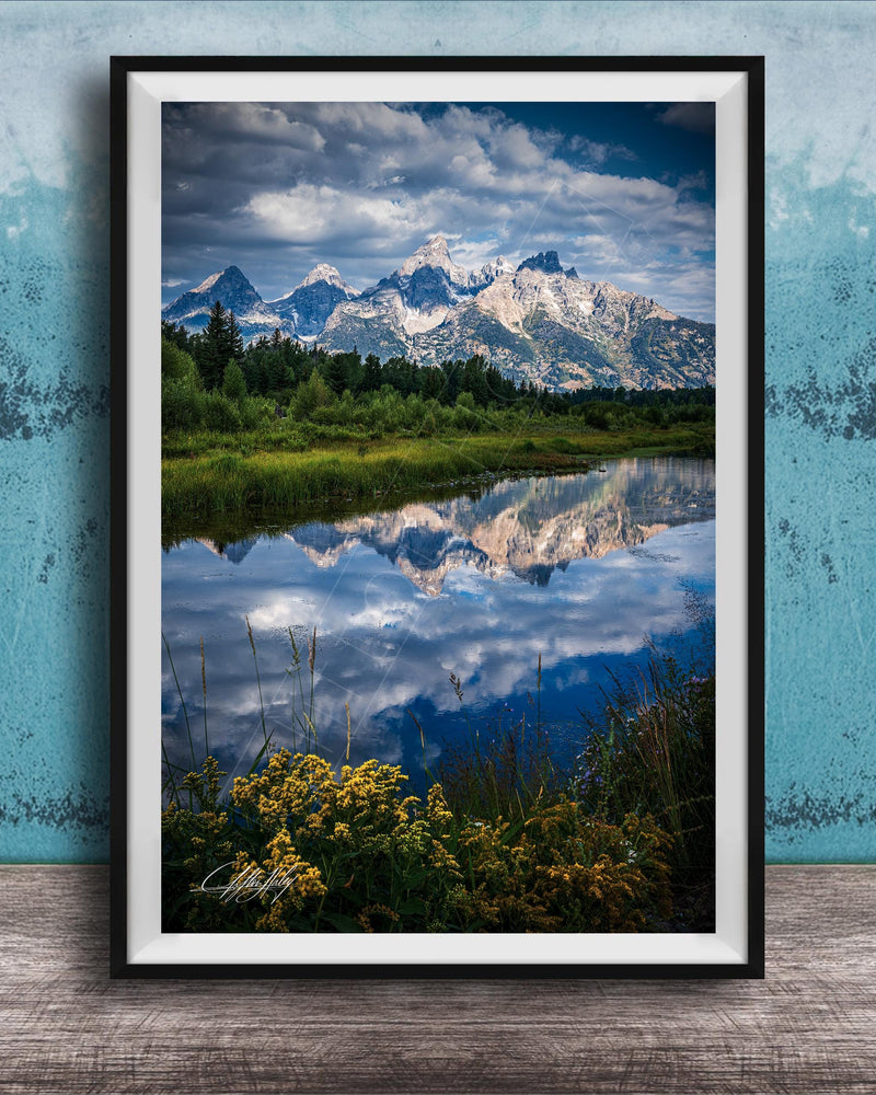 A framed photograph of a serene landscape with a mountain range reflected in a calm lake, surrounded by lush greenery and vibrant yellow flowers.
