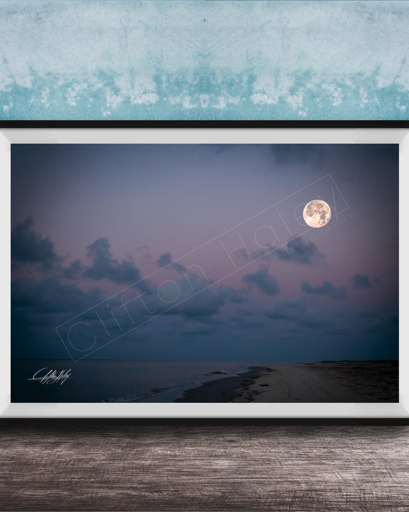 A framed photograph of a serene beach at night, with a full moon rising over the ocean and a cloudy sky.