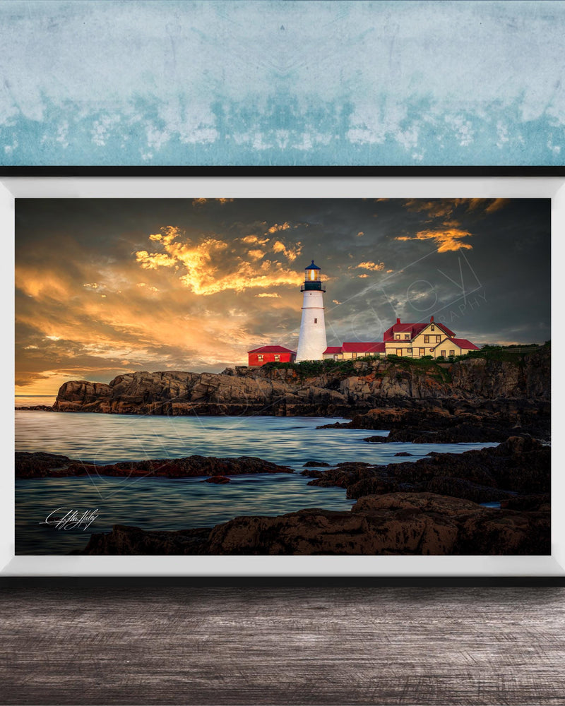 A framed photograph depicting a lighthouse on a rocky coastline at sunset, with the sky filled with clouds and the ocean in the background.
