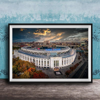 The image is a framed photograph of the New York Yankees stadium, which is a large, circular stadium located in the city of New York. The photograph captures the stadium's distinctive architecture and surrounding cityscape, with a dramatic sky in the background.