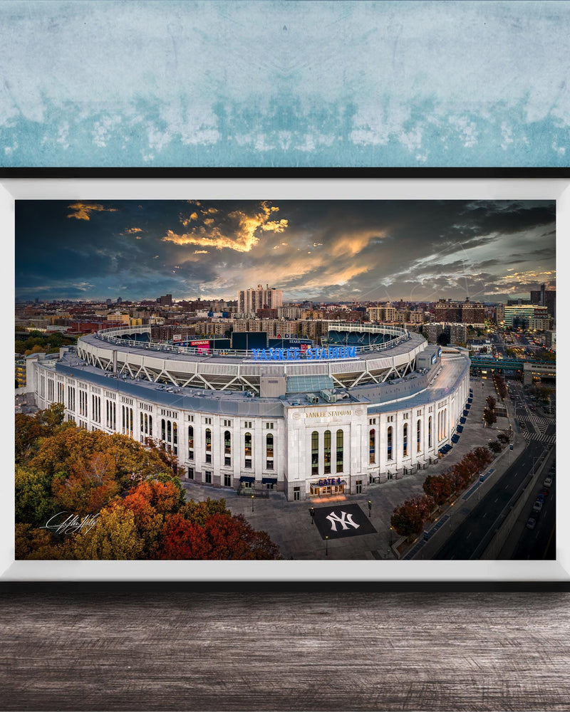 The image is a framed photograph of the New York Yankees stadium, which is a large, circular stadium located in the city of New York. The photograph captures the stadium's distinctive architecture and surrounding cityscape, with a dramatic sky in the background.