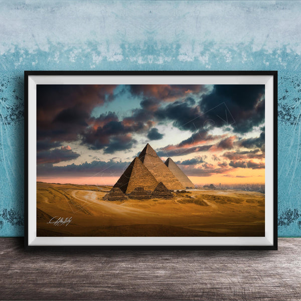 A framed photograph of the iconic pyramids of Giza against a dramatic sky at sunset, with the sun setting behind the sand dunes.