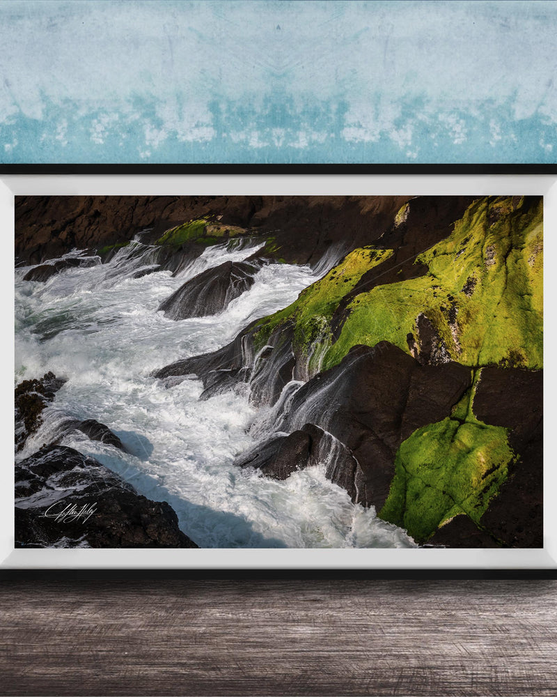 A framed photograph depicting a rocky coastline with flowing water and patches of green moss.