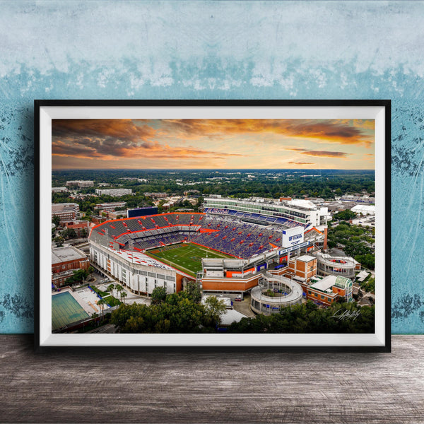 The image is a framed photograph of a large sports stadium, likely a football stadium, with a colorful sunset sky in the background.