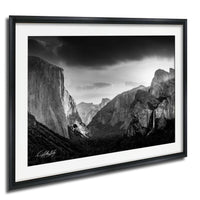 A black and white photograph of the majestic Yosemite Valley, with towering cliffs and a dramatic sky in the background.