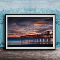 A framed photograph of a wooden pier extending into the ocean at sunset, with the sky filled with vibrant colors and clouds.