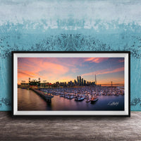 A framed photograph depicting a city skyline at sunset, with boats docked in a harbor and a large stadium in the background.