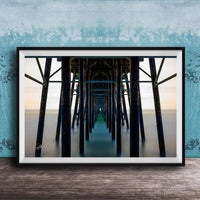 The image is a framed photograph of a long pier with multiple wooden columns extending into the distance, creating a sense of depth and perspective.