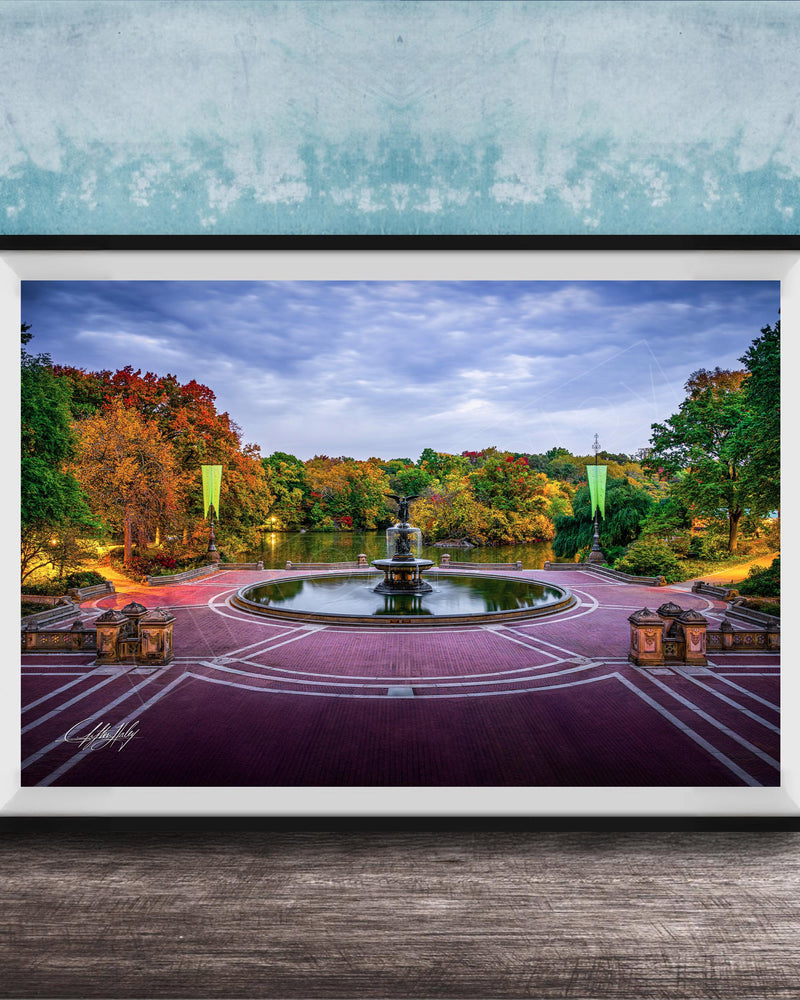 A framed photograph of a park with a fountain, surrounded by trees with autumn foliage, set against a blue wall.