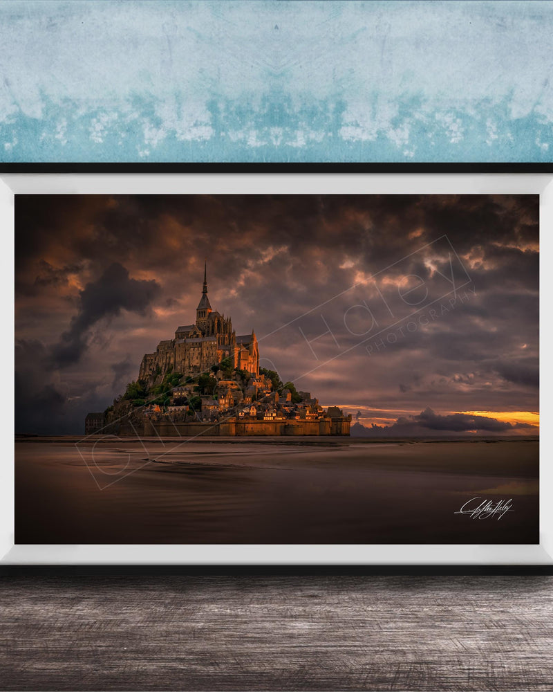 A framed photograph of the iconic Mont Saint-Michel in Normandy, France, with a dramatic sky in the background.