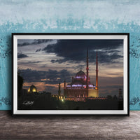 A framed photograph of a mosque at night, with its illuminated domes and minarets standing against a dark sky with clouds.