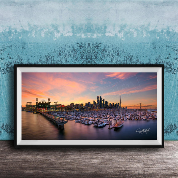 A framed photograph depicting a city skyline at sunset, with boats docked in a harbor and a large stadium in the background.