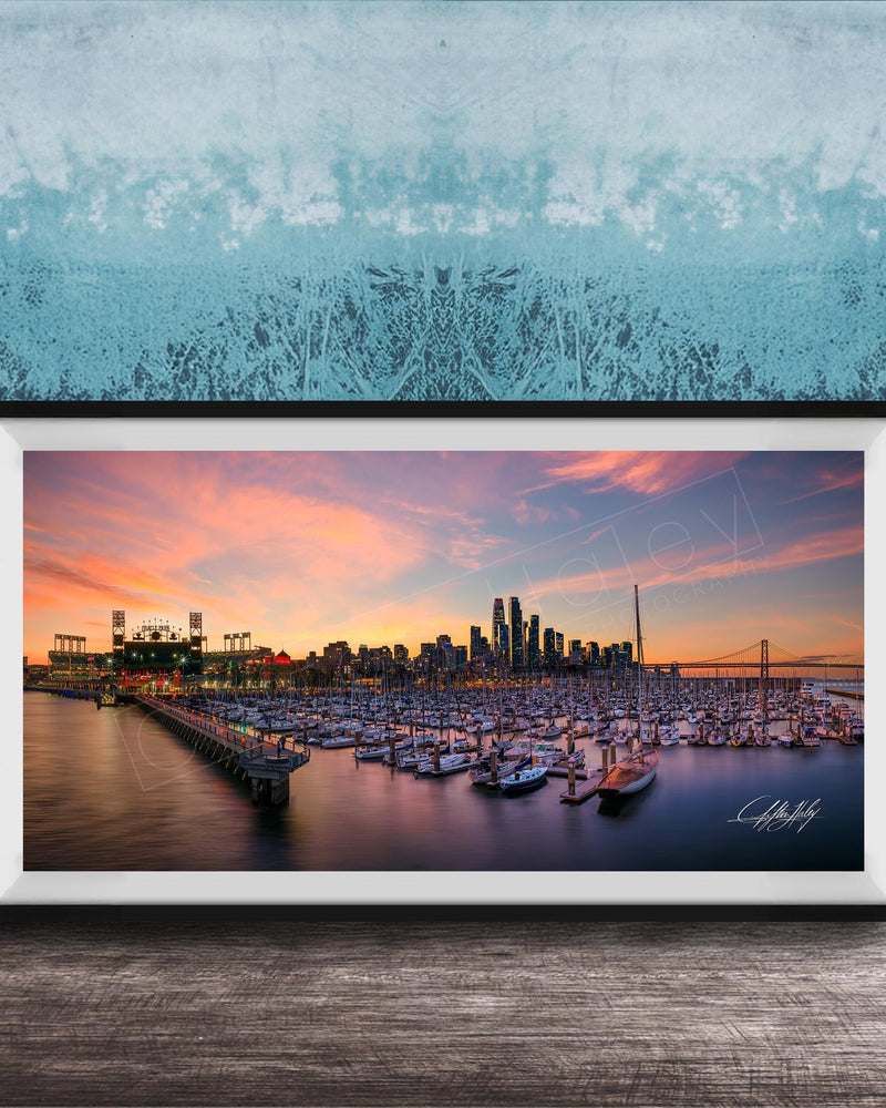 A framed photograph depicting a city skyline at sunset, with boats docked in a harbor and a large stadium in the background.