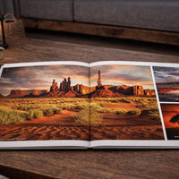 An open book displaying a collage of desert landscape photographs, including a desert canyon, a sunset, and a tree, with a candle and a mug of coffee on a wooden table in the background.