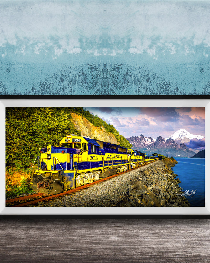 A framed photograph of a yellow and blue train traveling along a track through a mountainous landscape with a body of water in the background.