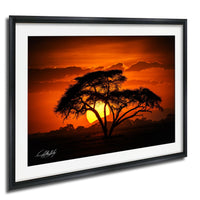 A framed photograph depicting a silhouetted tree against a vibrant orange and yellow sunset sky.