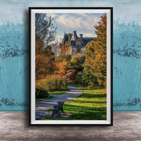 A framed photograph of a park with a winding path, a bench, and trees with autumn foliage. In the background, there is a large, ornate building with a gabled roof and multiple chimneys.