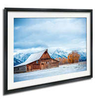 A rustic wooden barn with a sloping roof stands in a snowy landscape, with snow-capped mountains in the background.