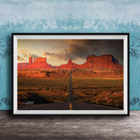 A framed photograph depicting a desert landscape with a winding road leading towards a series of red rock formations in the distance, set against a cloudy sky.