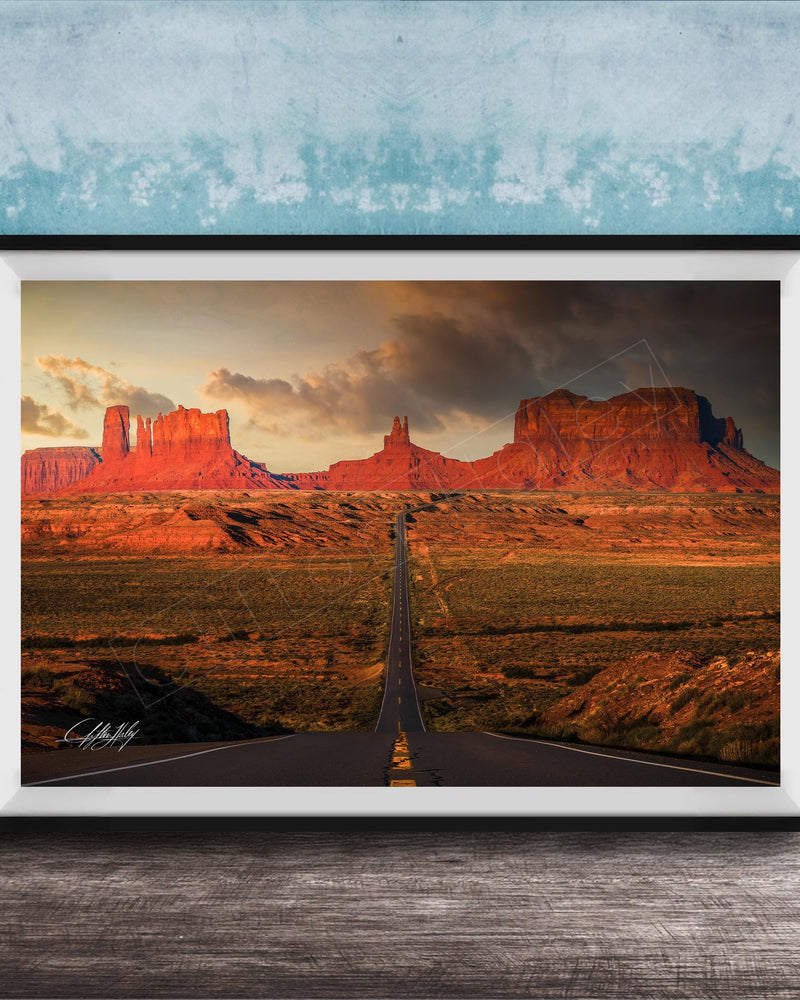 A framed photograph depicting a desert landscape with a winding road leading towards a series of red rock formations in the distance, set against a cloudy sky.